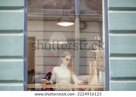 Young adult woman is reading magazine in cafe and drinking hot drink. Girl behind glass in cafe