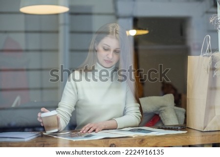 Young adult woman is reading magazine in cafe and drinking hot drink. Girl behind glass in cafe