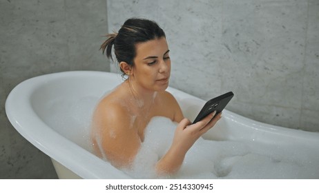 A young adult woman relaxes in a bubble bath at home, looking at her smartphone in a serene bathroom setting. - Powered by Shutterstock