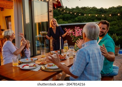 Young Adult Woman Serving Food To Her Husband And Parents Who Is Sitting At The Dining Table In The Backyard