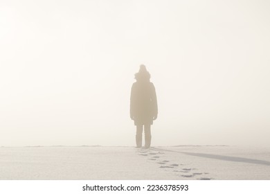 Young adult woman silhouette standing in nature mist and looking far away. Thinking about life. Back view. Fresh human boots footprints in fresh deep snow. Cold snowy winter day. 