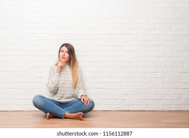 Young Adult Woman Sitting On The Floor Over White Brick Wall At Home With Hand On Chin Thinking About Question, Pensive Expression. Smiling With Thoughtful Face. Doubt Concept.