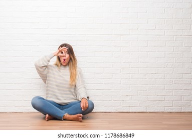 Young adult woman sitting on the floor over white brick wall at home doing ok gesture shocked with surprised face, eye looking through fingers. Unbelieving expression. - Shutterstock ID 1178686849