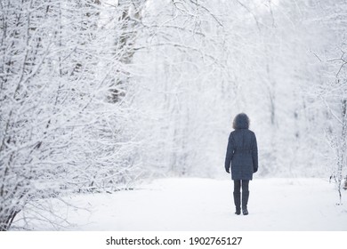 Young adult woman slowly walking on snowy trail at natural park in white winter day after blizzard. Fresh snow. Spending time alone in nature. Peaceful atmosphere. Back view.