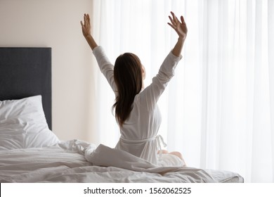 Young Adult Woman Wear Nightgown Raising Arms Waking Up Happy Concept In Early Good Morning Sitting In Cozy Bed, Fresh Rested Girl Stretch Awake After Healthy Sleep At Home Hotel Bedroom, Back View