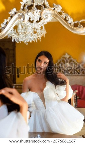 A young adult woman in a wedding dress preens herself in front of a mirror in a chic ancient castle.