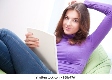 Young Adult Woman Writing In Her Copybook While Sitting On A Large Beanbag At Her Home