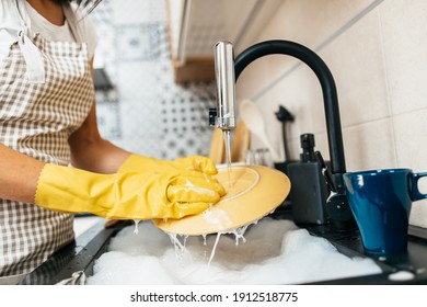 Young adult woman with yellow protective gloves washing her dishes on kitchen sink. Household and home hygiene routine.
