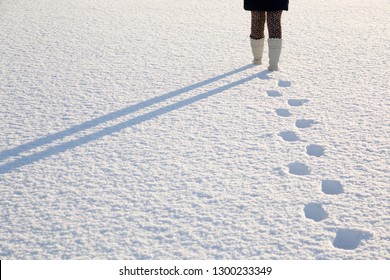 Young, adult woman's legs in white boots walking on fresh snow. Footprints behind human. Enjoying stroll in sunny, chilly winter day. Back view. Empty place for text, quote or sayings.