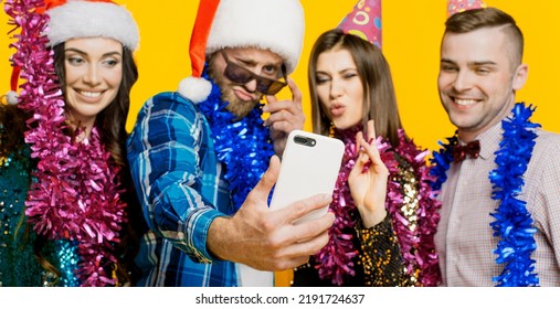 Young Adults 20s Wearing Party Hats And Santa Claus Caps And Tinsel Filming Video Or Taking Selfie On Smartphone For Social Media. Selective Focus On Smartphone In Hand