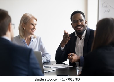 Young adults employees gather for briefing, during formal meeting multi ethnic people negotiating listening African skilled trainer participating at corporate workshop sit at desk in modern board room