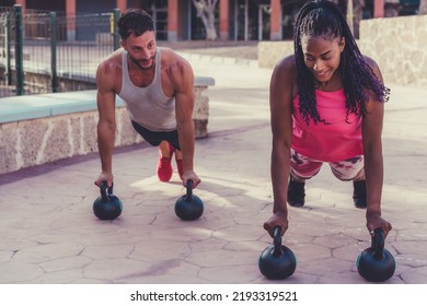 Young Adults Working Out Together Outside In The City. Man And Woman Holding A Plank Position And Smiling. Doing Plank On Kettlebell. Push-up On Weights. Fit Concept.