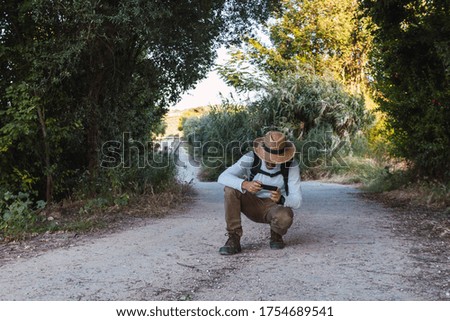 Young adventurer in hat sending messages with mobile phone in the field. Remote communication via mobile phone. Traveling alone without losing connection. Selective focus.