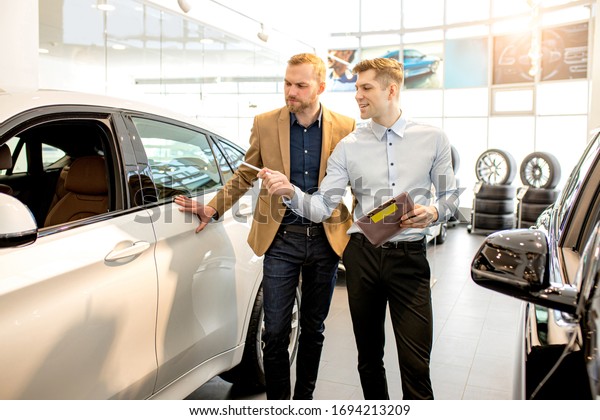 young affable consultant talking about\
features of new car to customer. business man listen to him,\
choosing new auto in\
dealership