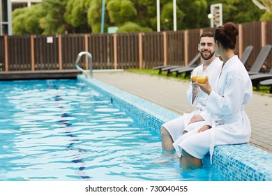 Young affectionate couple with drinks having talk while sitting by swimming pool with their legs in water