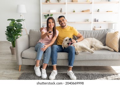 Young affectionate multiracial couple sitting on couch with pet dog and watching television. Diverse boyfriend and girlfriend spending time together, enjoying good movie at home