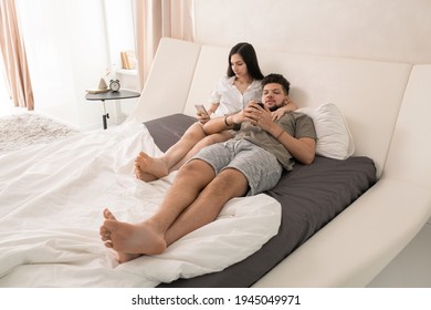 Young affectionate restful couple with smartphones relaxing on large double bed and scrolling through posts in social networks