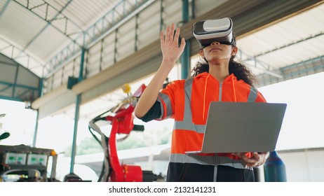 Young Africa American Engineer Wearing Virtual Reality Headset In The Robotic Arm Factory. Holding Laptop And See Demonstration Robotic Arm Via Computer Software. VR Technology Industrial Concept