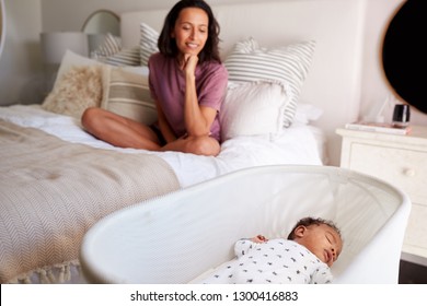 Young African American Adult Mother Sitting Cross Legged On Her Bed Looking Down At Her Three Month Old Baby Sleeping In His Cot, Close Up