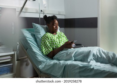 Young african american adult with sickness having smartphone in hand while laying in hospital ward. Sick teenager using internet technology for remote family communication and chatting - Powered by Shutterstock