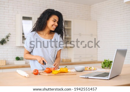 Young african american adult woman in striped T-shirt cooking food at home in kitchen using grey laptop to learn preparing a vegy pizza, chopping vegetables with knife, watches cooking channels