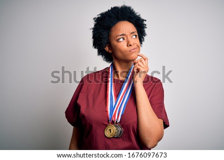 Young African American afro athlete woman with curly hair wearing medals for competition with hand on chin thinking about question, pensive expression. Smiling with thoughtful face. Doubt concept.