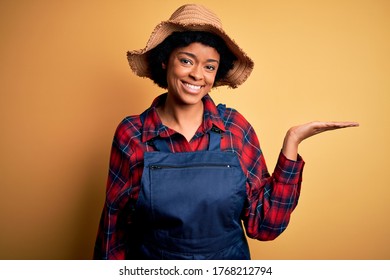 Young African American Afro Farmer Woman With Curly Hair Wearing Apron And Hat Smiling Cheerful Presenting And Pointing With Palm Of Hand Looking At The Camera.