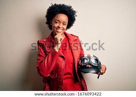 Young African American afro motorcyclist woman with curly hair holding motorcycle helmet with hand on chin thinking about question, pensive expression. Smiling and thoughtful face. Doubt concept.