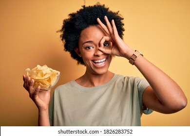 Young African American afro woman with curly hair holding bowl with chips potatoes with happy face smiling doing ok sign with hand on eye looking through fingers