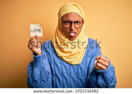 Young African American afro woman wearing muslim hijab holding love reminder paper message annoyed and frustrated shouting with anger, crazy and yelling with raised hand, anger concept