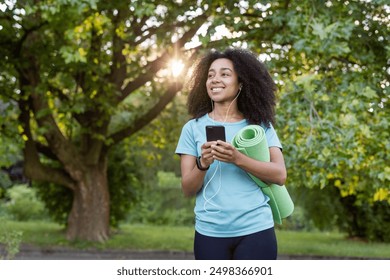 Young African American athlete carrying yoga mat, listening to music, smiling while using smartphone outdoors in park. Fitness enthusiast enjoying nature, technology, and healthy lifestyle. - Powered by Shutterstock