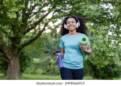 Young African American athlete walking in park holding yoga mat and water bottle. Wearing headphones and smiling confidently. Outdoor fitness and healthy lifestyle concept. - Powered by Shutterstock