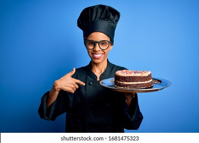 Young African American Baker Woman Wearing Cooker Uniform And Hat Holding Cake With Surprise Face Pointing Finger To Himself