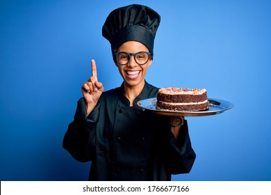 Young African American Baker Woman Wearing Cooker Uniform And Hat Holding Cake Surprised With An Idea Or Question Pointing Finger With Happy Face, Number One