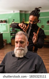 Young African American Barber And Dreadlocks, With Coronavirus Pandemic Prevention Mask, Combs With A Comb And His Hands A Mature White Caucasian Man With A White Gray Beard In A Barber Shop.  Hipster