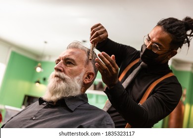 Young African American Barber And Dreadlocks, With Coronavirus Pandemic Prevention Mask, Combs With A Comb And His Hands A Mature White Caucasian Man With A White Gray Beard In A Barber Shop.  Hipster