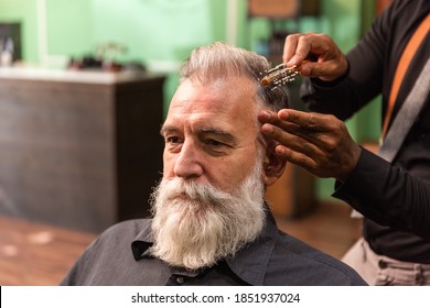 Young African American Barber And Dreadlocks, With Coronavirus Pandemic Prevention Mask, Combs With A Comb And His Hands A Mature White Caucasian Man With A White Gray Beard In A Barber Shop. 