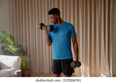 Young African American Black Male Working Out In Living Room At Home Doing A Bicep Curl With Heavy Dumbbell Weights. Keeping Fit And Healthy With Strength Training