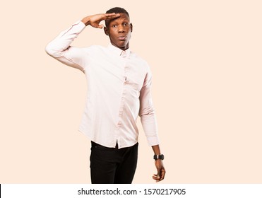 Young African American Black Man Greeting The Camera With A Military Salute In An Act Of Honor And Patriotism, Showing Respect Against Beige Wall