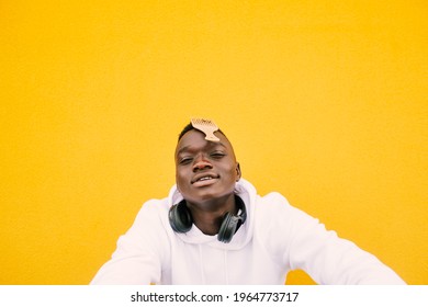 Young african American black man combing his afro hair while seated on a yellow wall background, wearing a white sweatshirt and wireless headphones. Looking to the camera. Copy space
