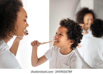 A young African American boy, with curly hair and a white t-shirt, smiles at a woman while learning to brush his teeth. She is demonstrating the proper technique with her own toothbrush - Powered by Shutterstock