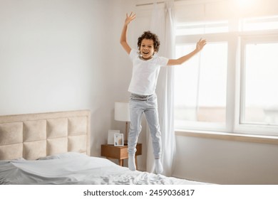 A young African American boy with curly hair is captured mid-jump on a bed in a well-lit bedroom, his face expressing pure joy and exhilaration - Powered by Shutterstock