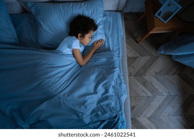 A young African American boy is laying comfortably in bed with a soft blue comforter covering him. He appears relaxed and at ease, possibly getting ready to sleep or just waking up, view above - Powered by Shutterstock