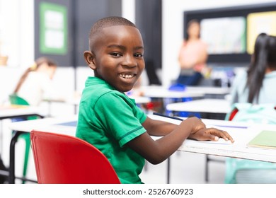 Young African American boy sits at classroom desk in school, smiling at camera. Behind him, middle-aged biracial woman teaching, with recycle poster visible, learning, education, unaltered - Powered by Shutterstock