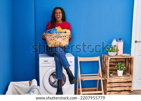 Young african american with braids holding laundry basket sitting on washing machine angry and mad screaming frustrated and furious, shouting with anger. rage and aggressive concept. 