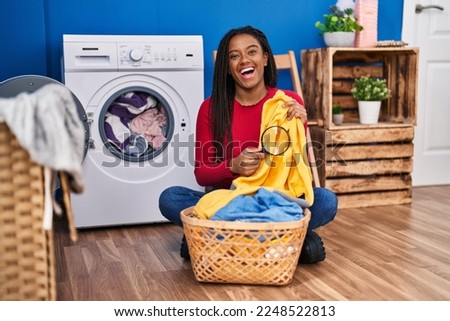 Young african american with braids holding magnifying glass looking for stain at clothes smiling and laughing hard out loud because funny crazy joke. 