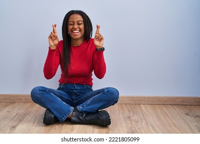 Young african american with braids sitting on the floor at home gesturing finger crossed smiling with hope and eyes closed. luck and superstitious concept. 