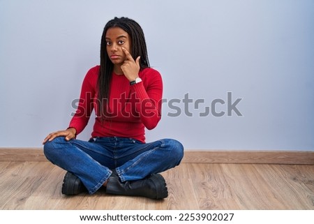 Young african american with braids sitting on the floor at home pointing to the eye watching you gesture, suspicious expression 