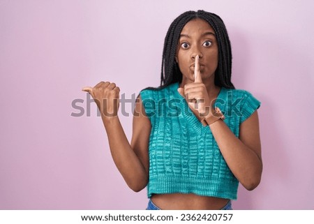 Young african american with braids standing over pink background asking to be quiet with finger on lips pointing with hand to the side. silence and secret concept. 