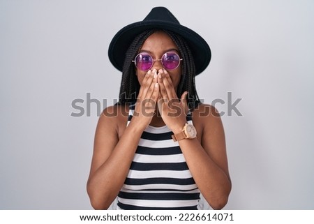 Young african american with braids wearing hat and sunglasses laughing and embarrassed giggle covering mouth with hands, gossip and scandal concept  Foto d'archivio © 